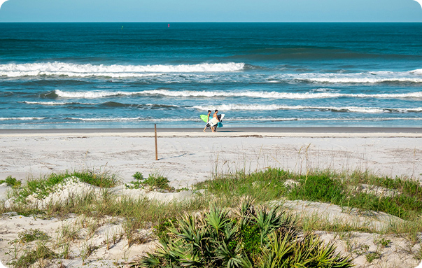 Two surfers carrying boards across the sand toward the water at New Smyrna Beach, Florida